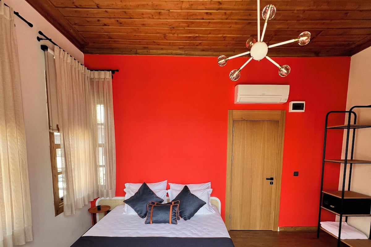 A room with a wooden ceiling, a bed with white and dark bedding, a bunk bed, a ceiling fan, and a window with curtains.