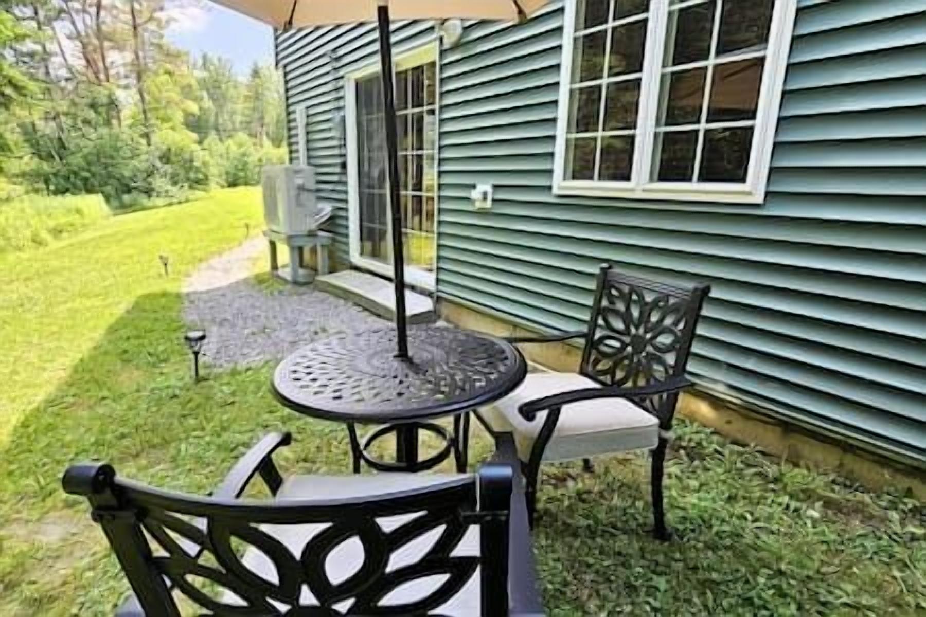 A patio with a round table and two chairs, a small garden area, and a house with green siding and a window.