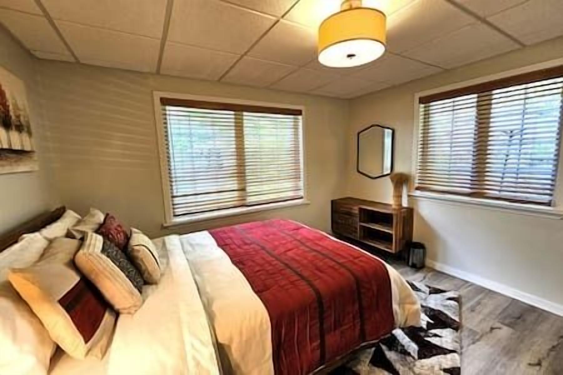 Ein Schlafzimmer mit einem Bett, zwei Fenstern mit Jalousien, einer Holzkommode und einem Spiegel an der Wand.