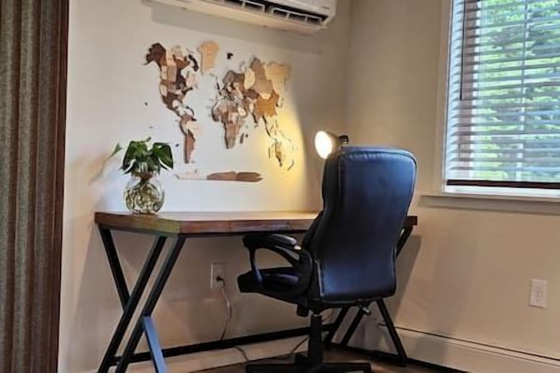 Ein Zimmer mit Schreibtisch, Stuhl, einer Weltkarte als Wandbild, einer Topfpflanze und einem Fenster mit Jalousien.