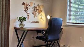 Ein Zimmer mit Schreibtisch, Stuhl, einer Weltkarte als Wandbild, einer Topfpflanze und einem Fenster mit Jalousien.