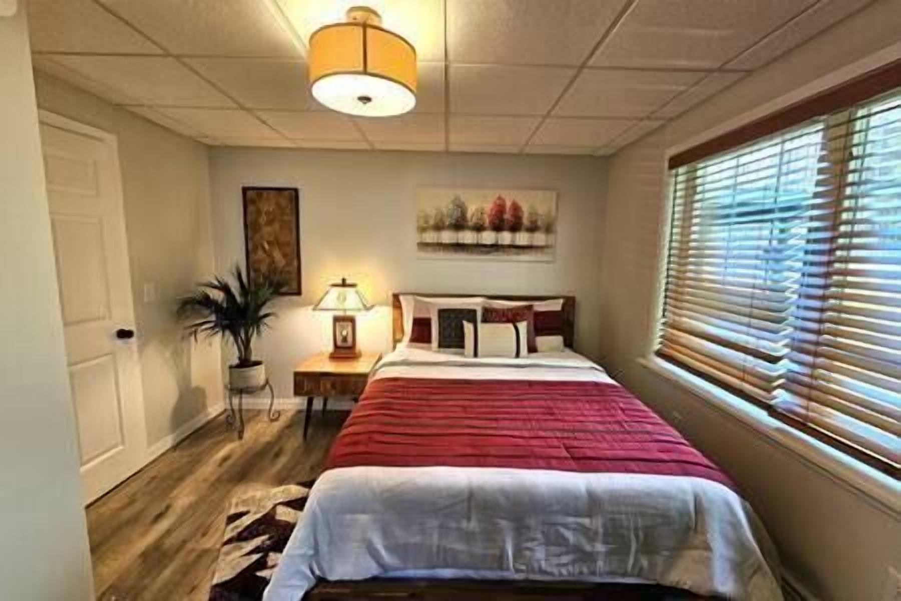 Un dormitorio con cama, mesita de noche, lámpara, ventana con persianas y puerta.