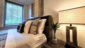 A hotel room with a large bed, a nightstand with a lamp, a potted plant, and a window with blinds.
