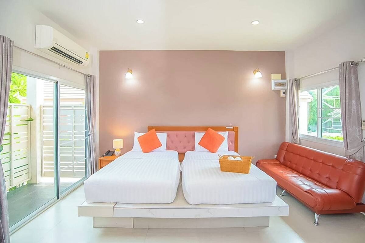 A double bed with white linens and orange pillows, a red leather sofa, and a small table with a basket.