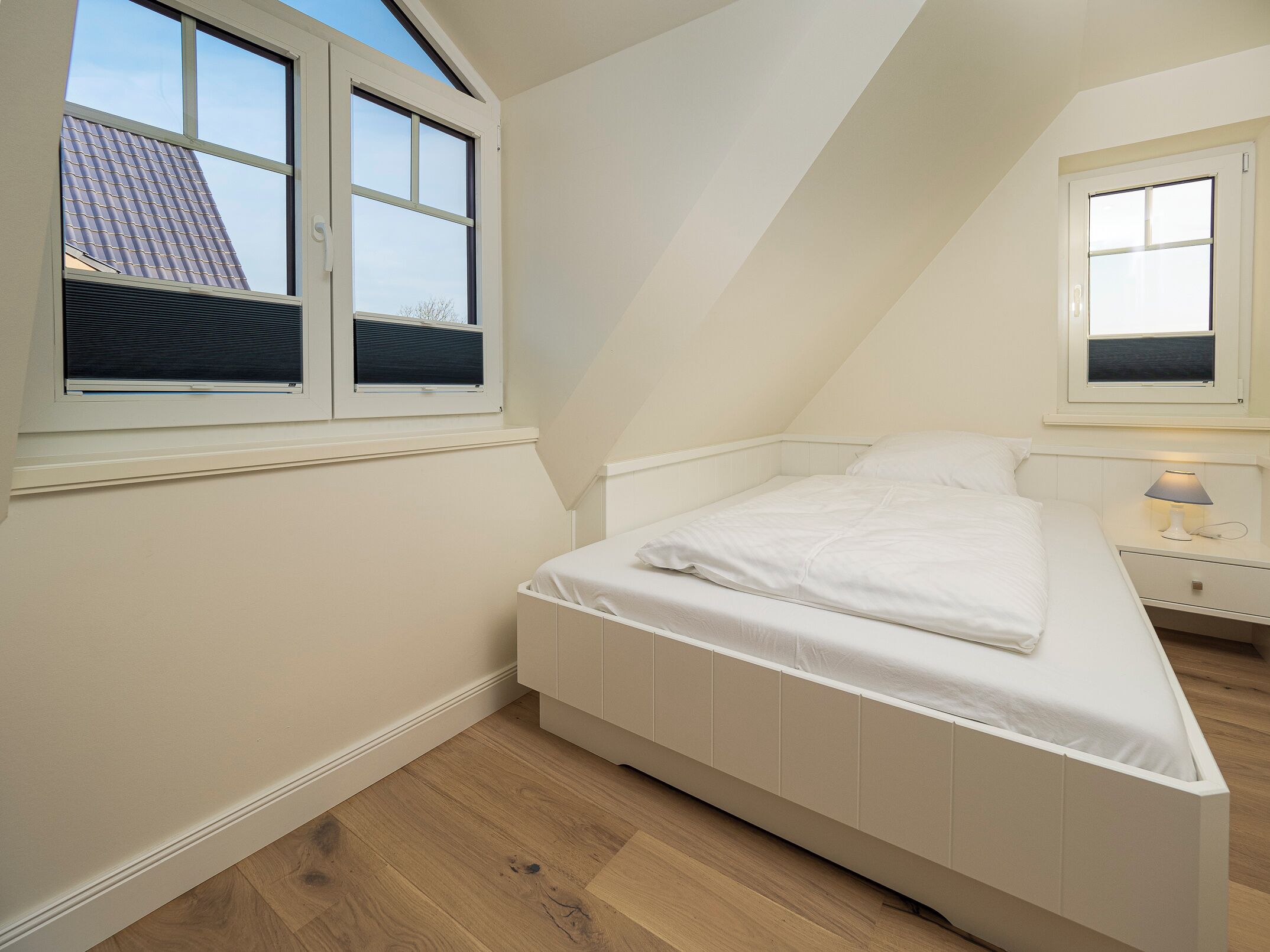 A bedroom with a sloped ceiling, a bed, a nightstand with a lamp, and two windows.