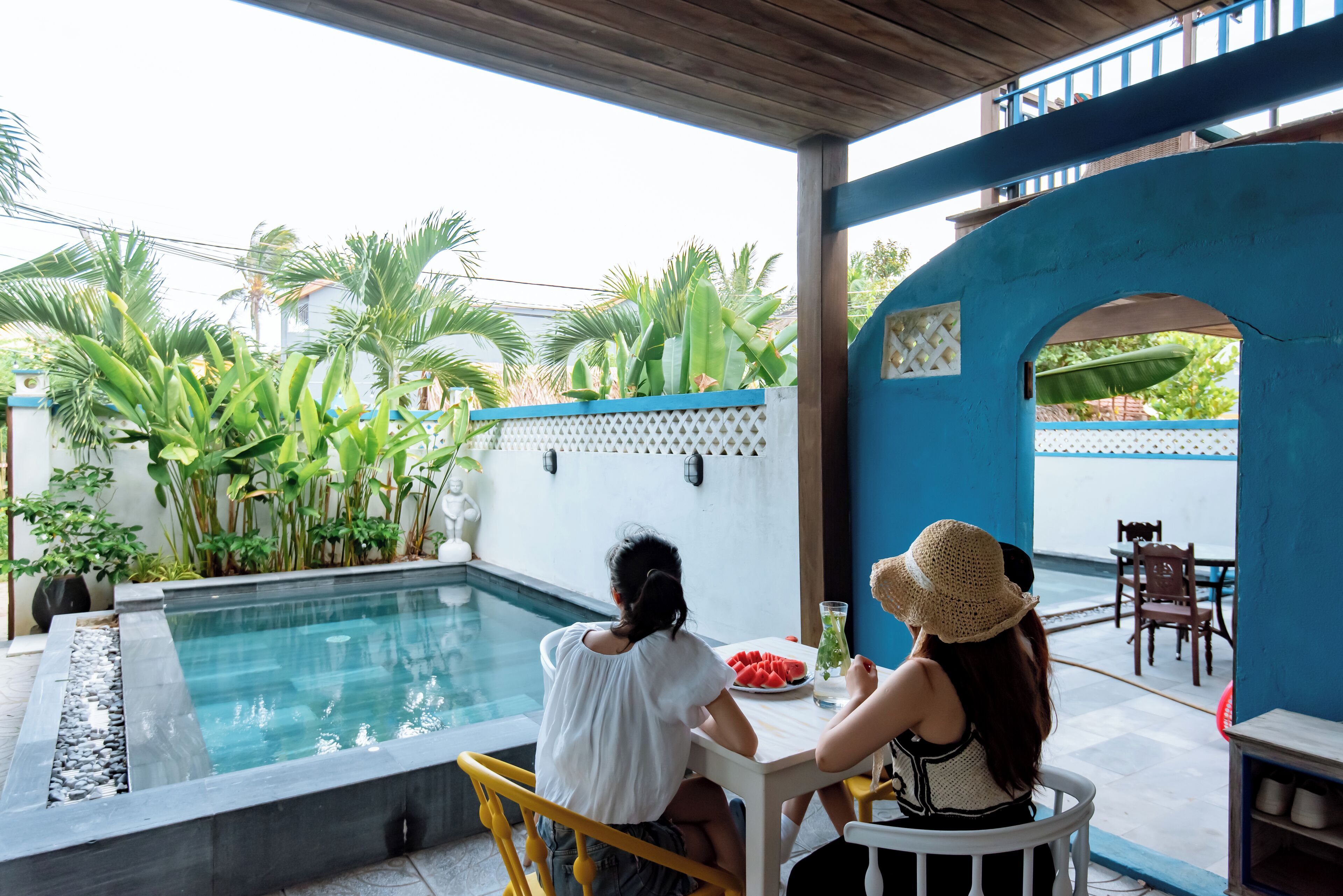 Un espace aménagé au bord de la piscine, avec une table et des chaises, un mur bleu et des plantes tropicales.