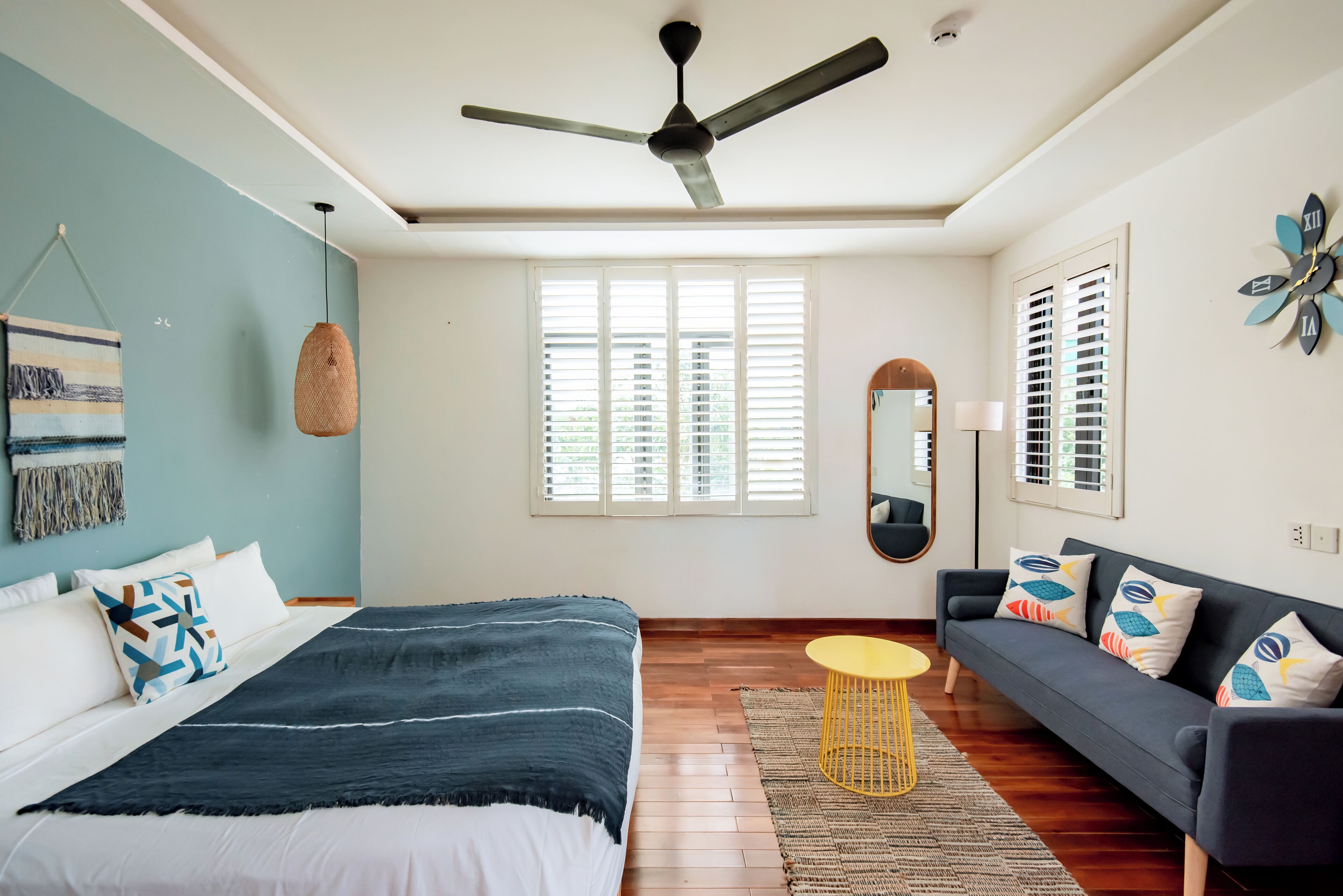 A bedroom with a bed, a sofa, a ceiling fan, a window with blinds, and a wall clock.