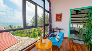 Un balcón con vista al océano, una silla azul, una mesita con un florero y una obra de arte roja y dorada en la pared.