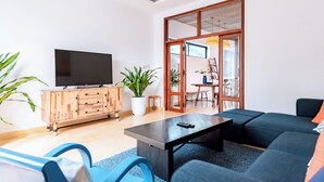 Una sala de estar moderna con televisor de pantalla plana, mueble de madera, sofá azul y un área de comedor visible a través de una puerta de vidrio.