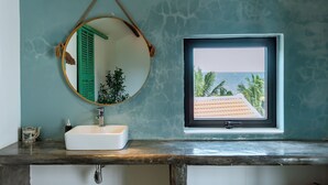 Un baño con un lavabo blanco, un espejo redondo y una ventana con vistas a palmeras y una playa.