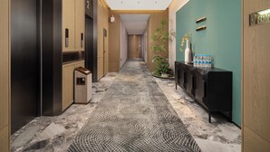 A modern hallway with a patterned carpet, wooden and black elevator doors, a teal accent wall, and a water dispenser on a console. - Meihao Lizhi Hotel (Xi'an Lintong Huaqingchi Subway Station Branch) (Xi'an)