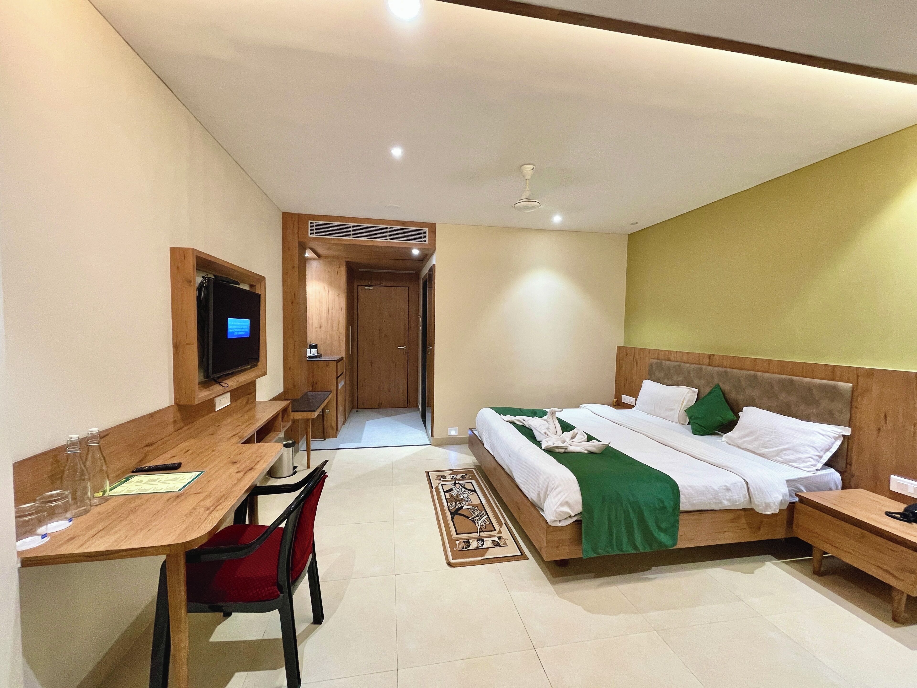 A hotel room with a wooden desk, a bed with green and white bedding, a TV mounted on the wall, and a ceiling fan.