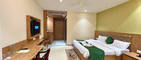 A hotel room with a wooden desk, a bed with green and white bedding, a TV mounted on the wall, and a ceiling fan.