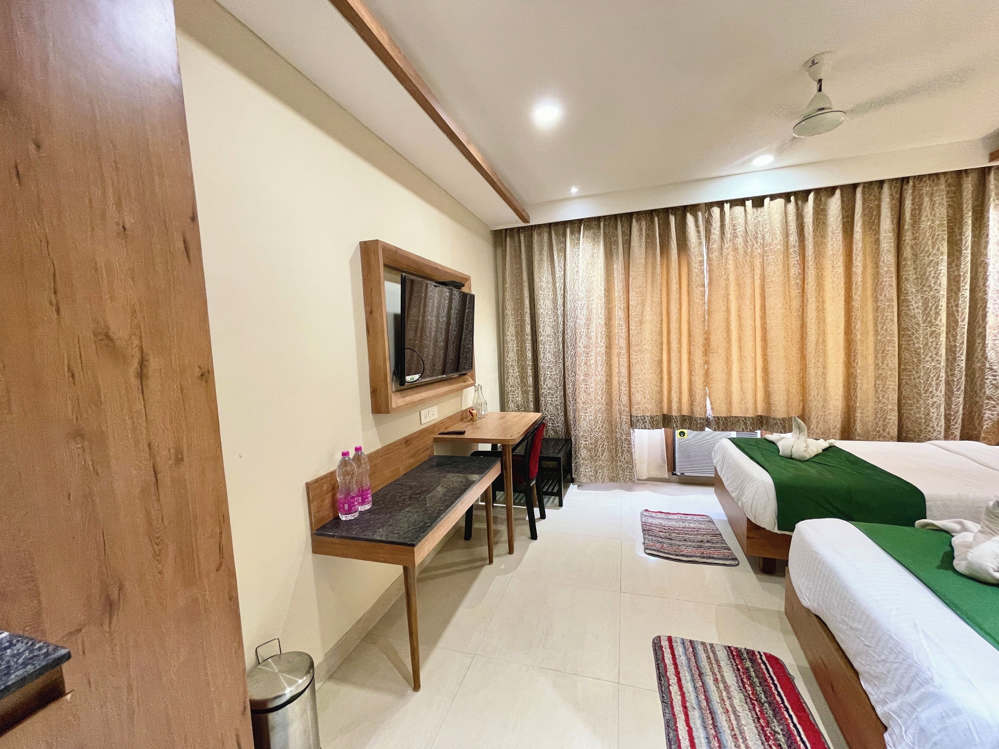 A hotel room with a bed, a desk with a chair, a TV mounted on the wall, and a window with curtains.