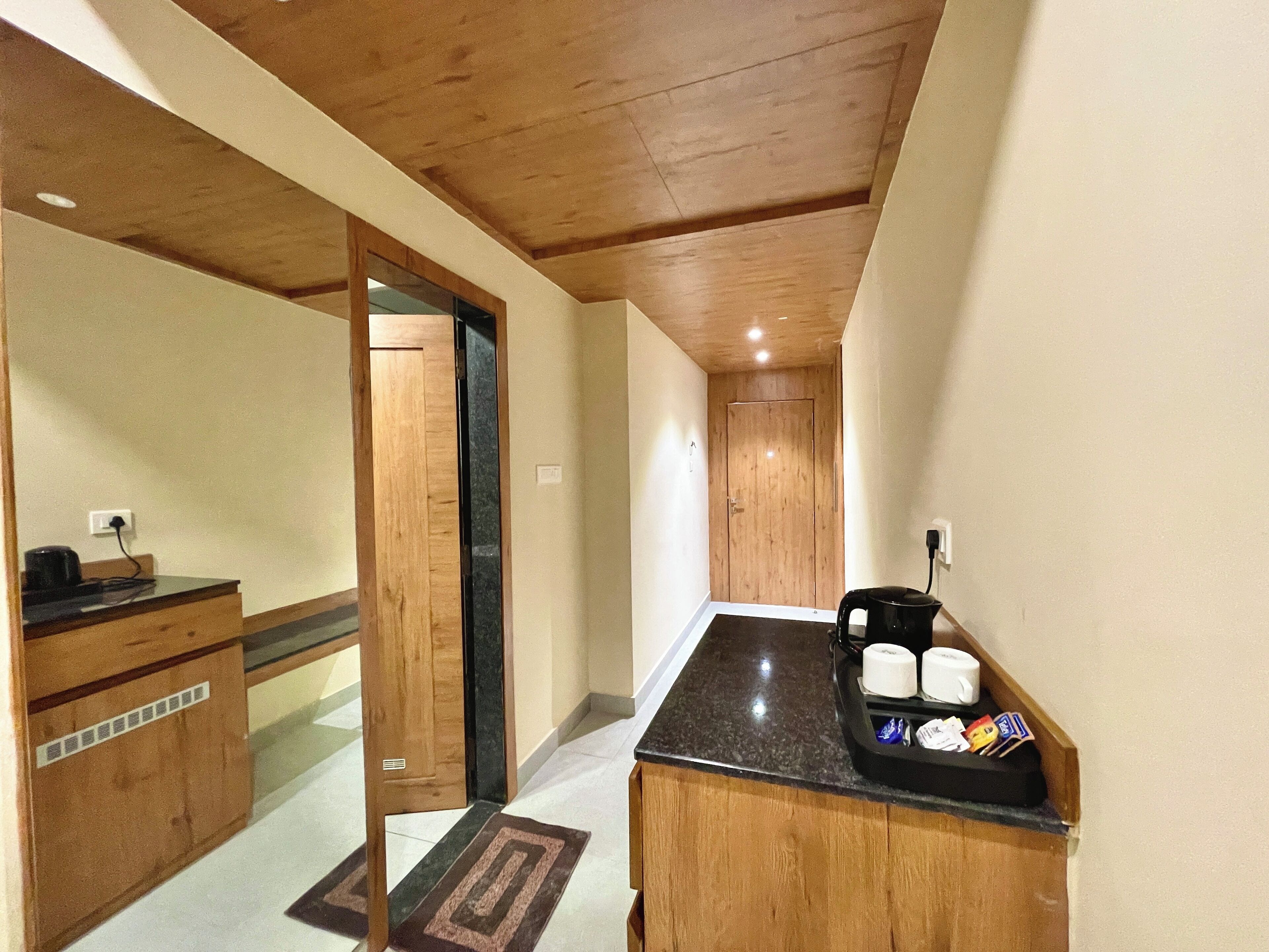Un pasillo estrecho con suelo y techo de madera. Se observa una pequeña área de kitchenette con una encimera, una tetera y algunos alimentos envasados. Una puerta da a otra habitación.