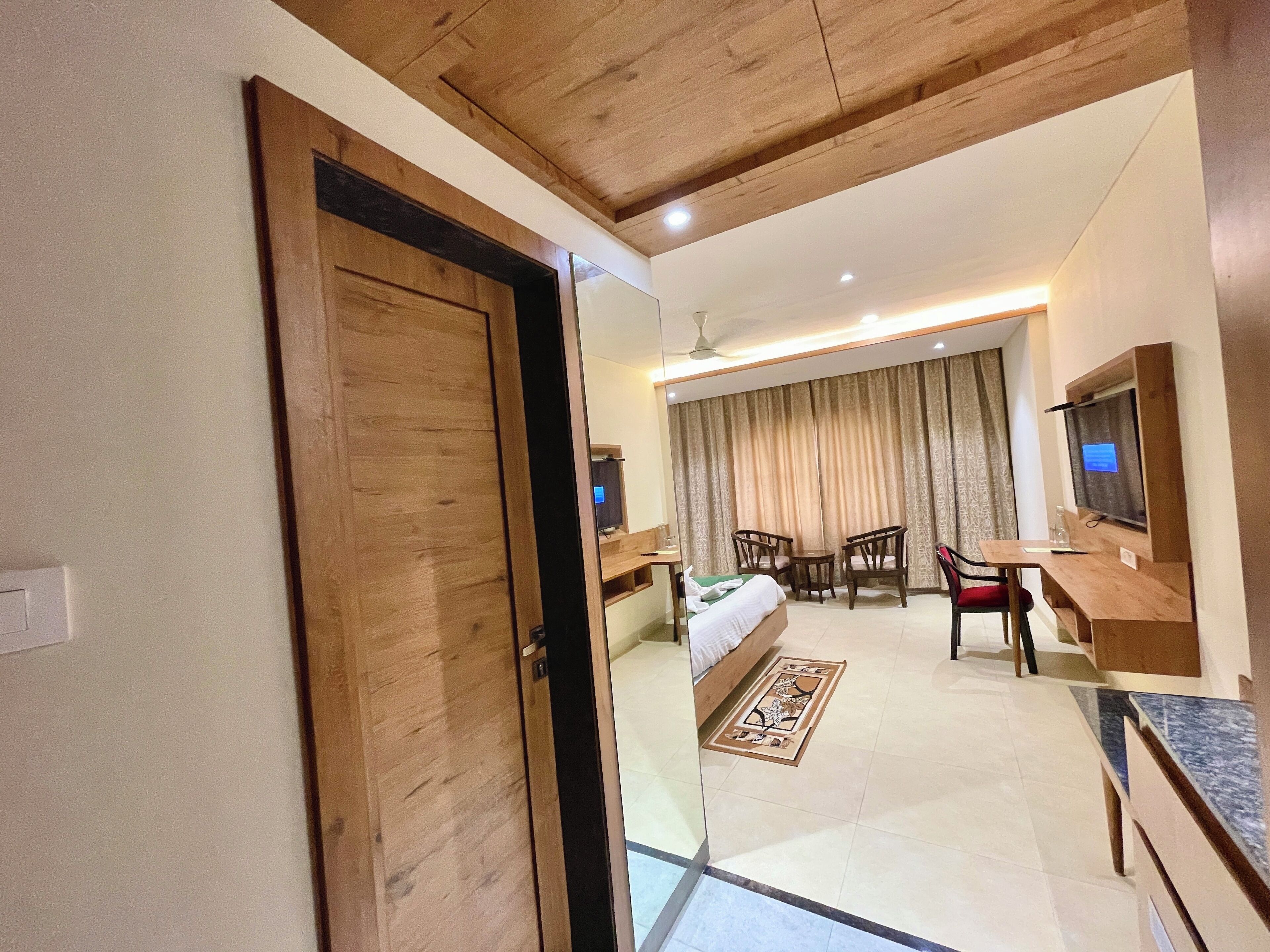 A hotel room with a wooden door, a bed with a white comforter, a desk with a chair, a TV, and a large window with curtains.