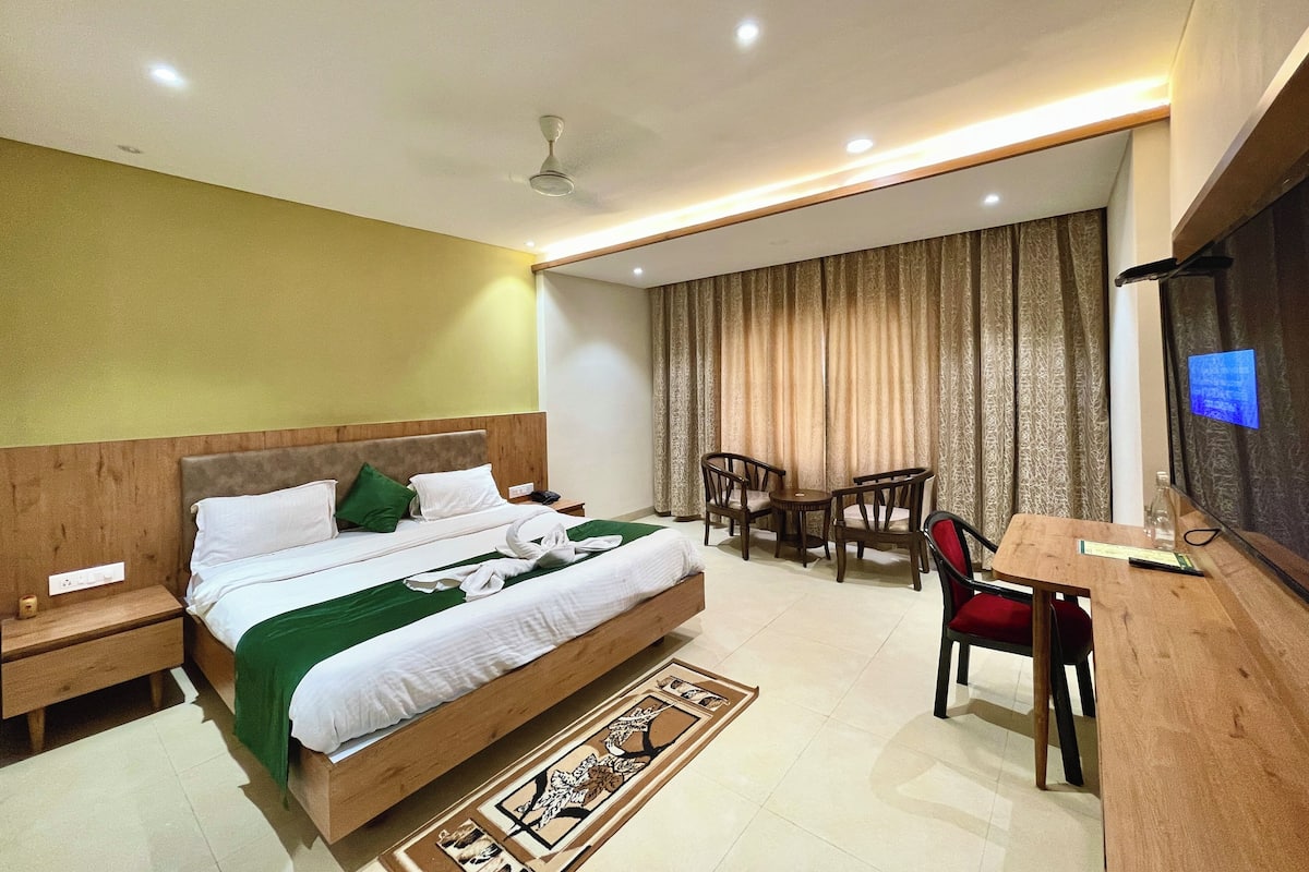 A hotel room with a bed, bedside tables, a desk with chairs, a television, and a large window with curtains.