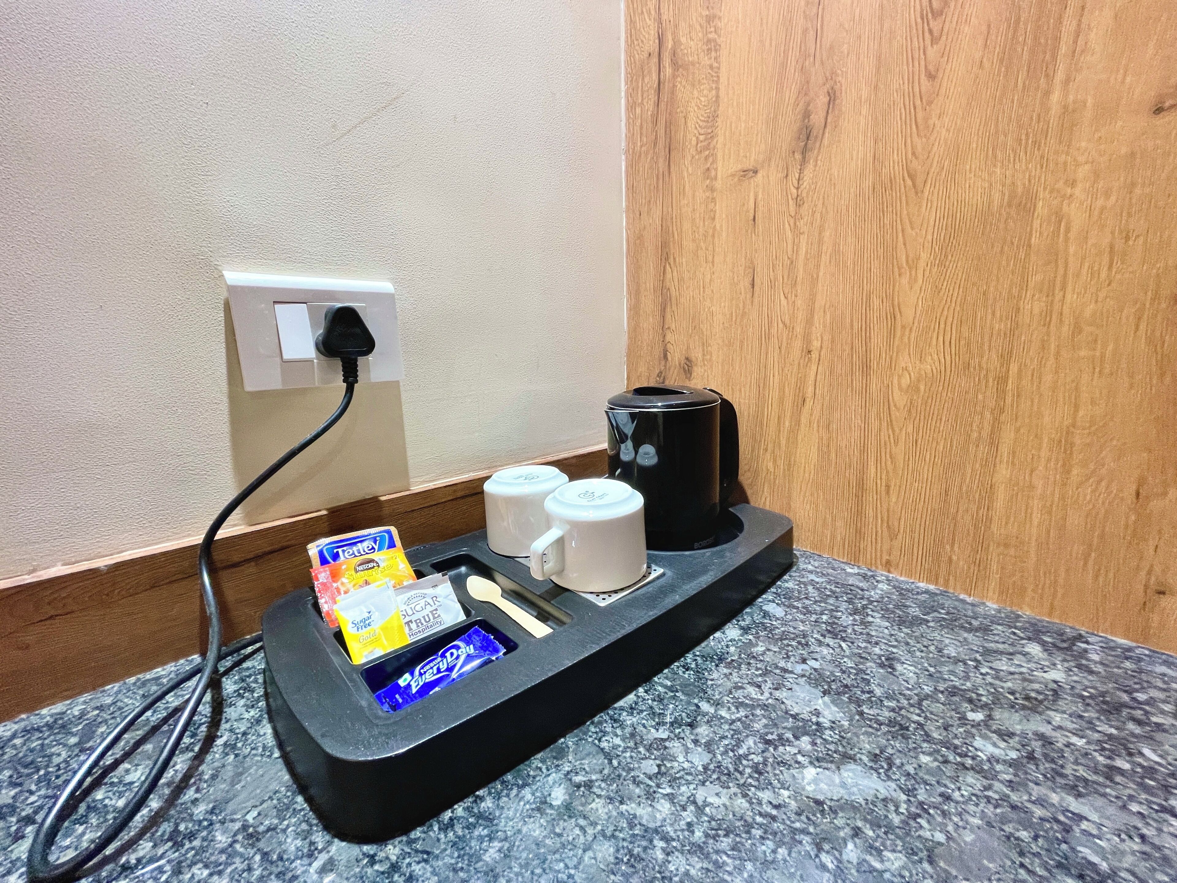 A countertop with a coffee maker, two white mugs, a black power strip, and a snack bar with sugar packets and a chocolate bar.