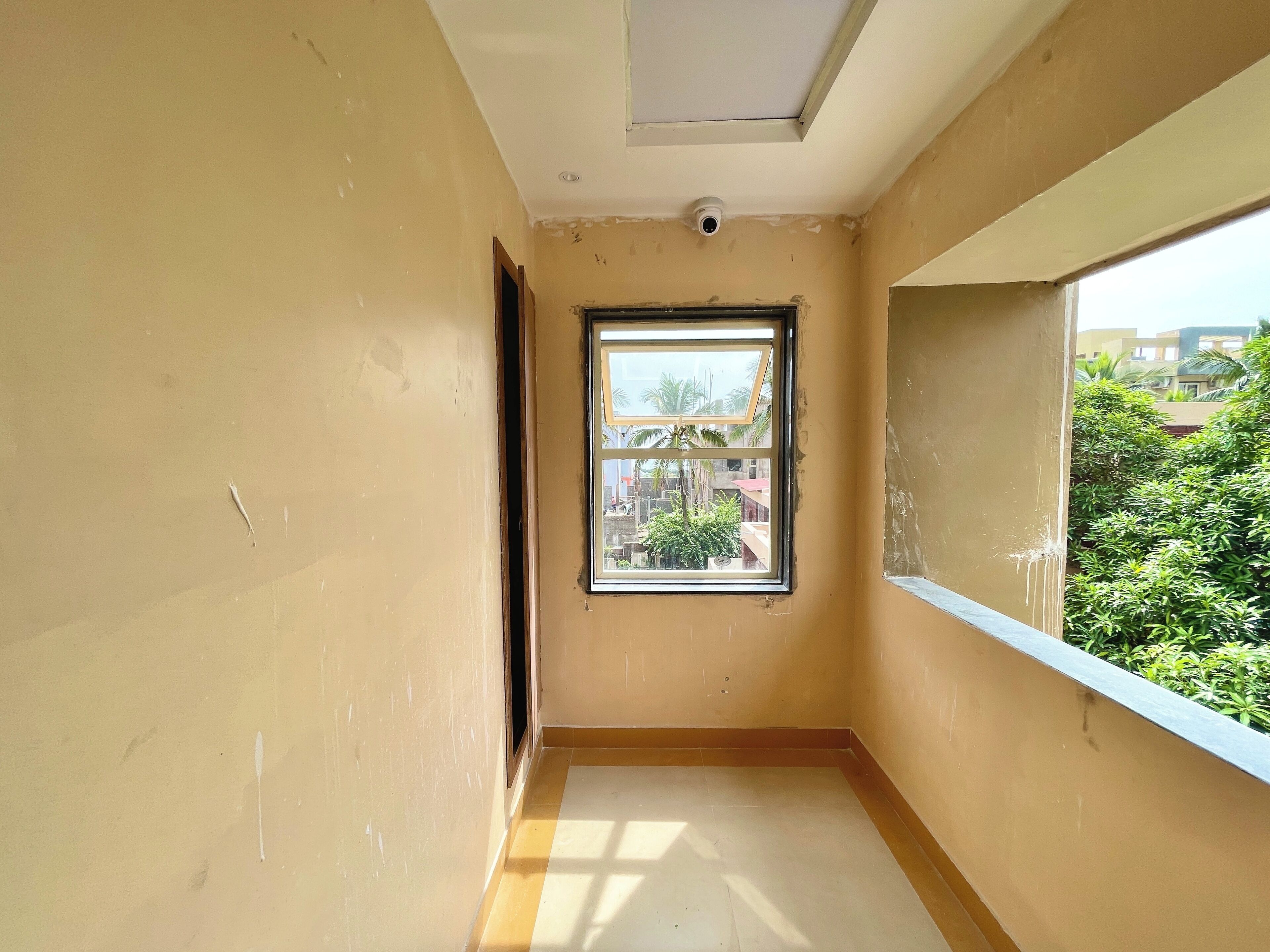 Un pasillo estrecho con una ventana, una cámara de seguridad en el techo y vistas a áreas verdes y edificios.