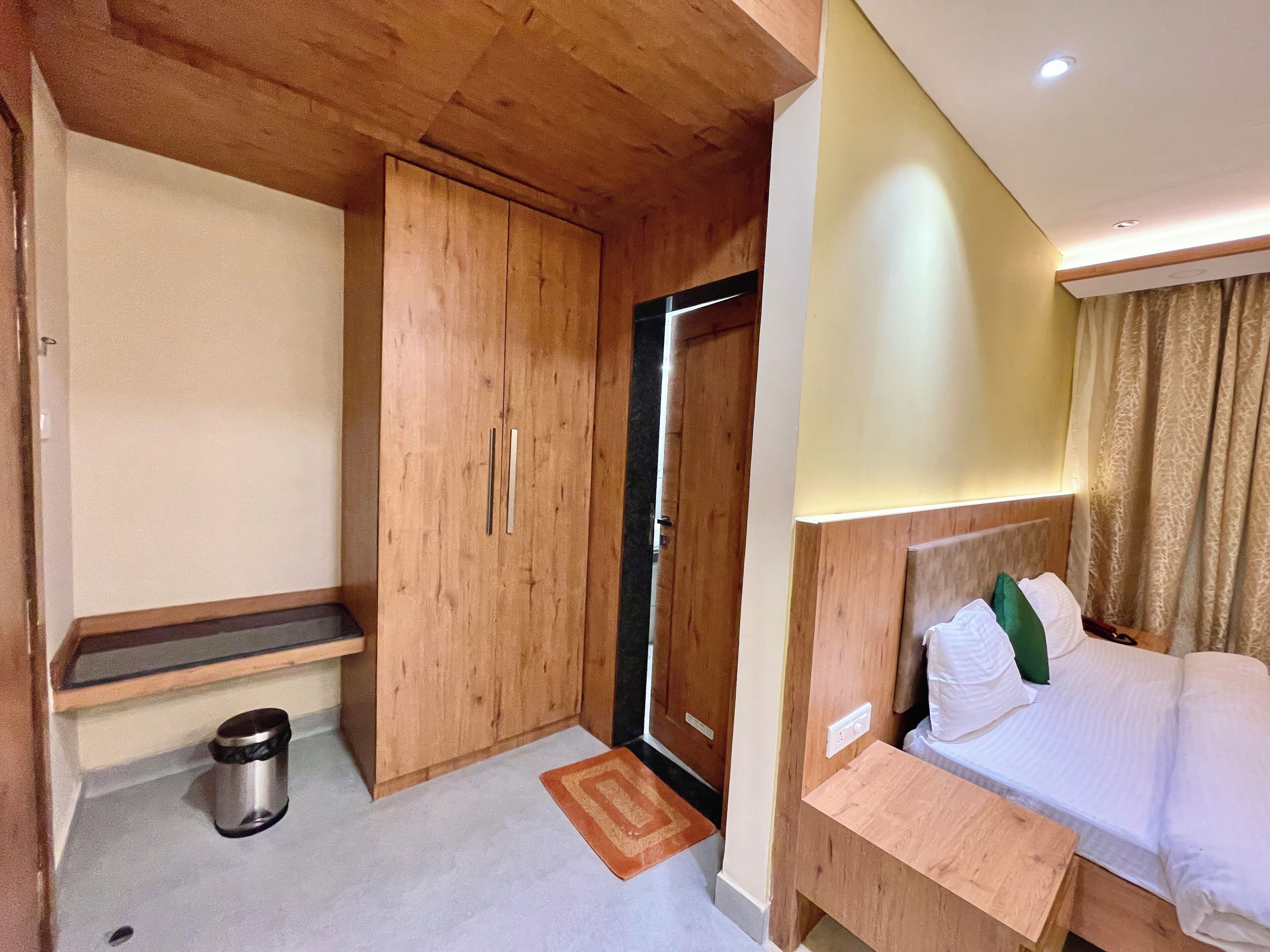 A hotel room with a wooden wardrobe, a bedside table, a bed with white bedding and green pillows, a small trash bin, and a door leading to another room.