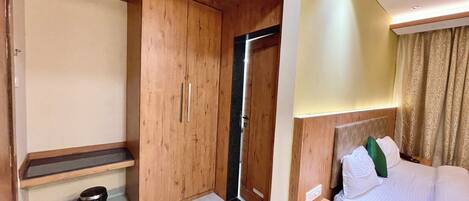 A hotel room with a wooden wardrobe, a bedside table, a bed with white bedding and green pillows, a small trash bin, and a door leading to another room.