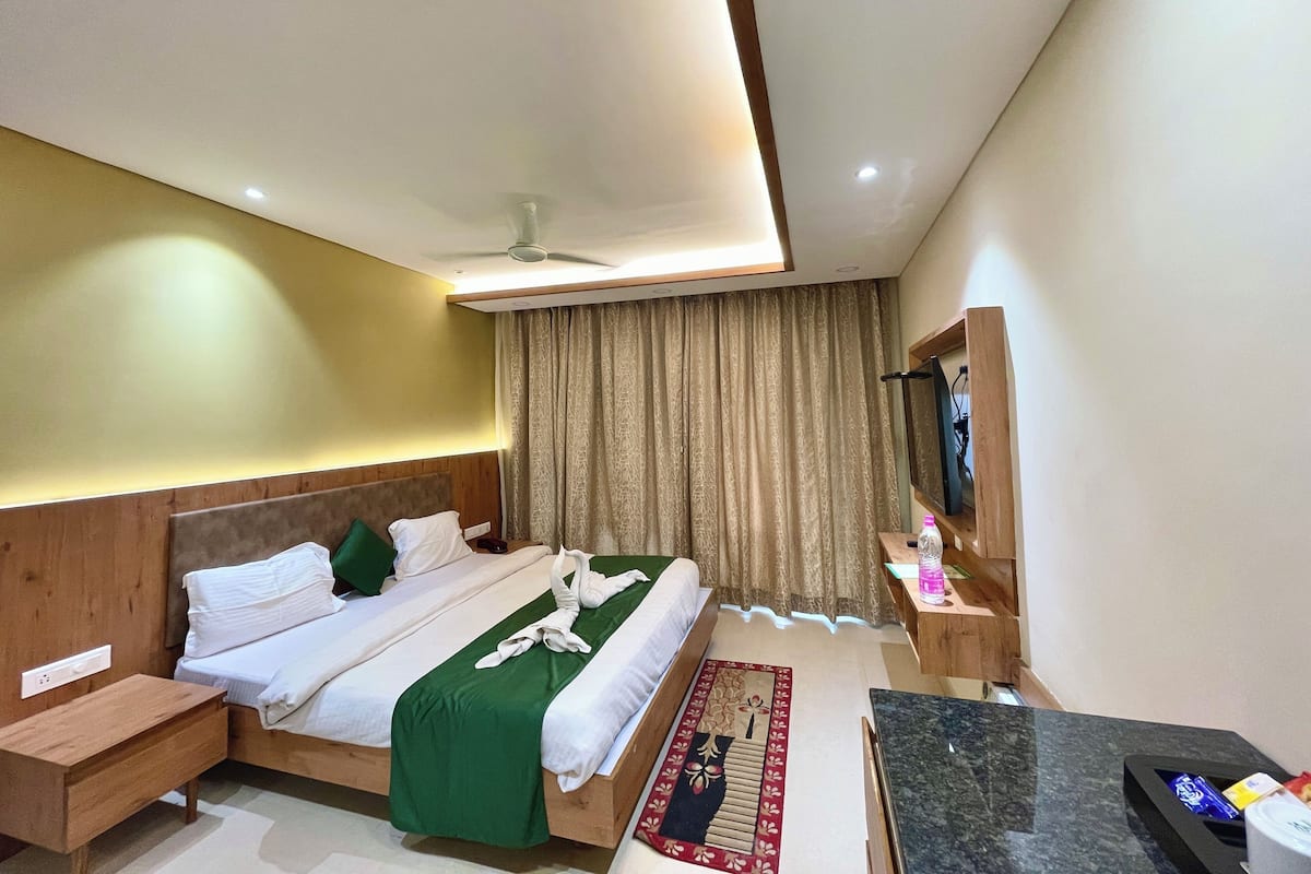 A modern hotel room with a wooden headboard, a bed with white linens, a green throw blanket, a small bedside table, a nightstand, a desk with a chair, a flat-screen TV, and a ceiling fan.