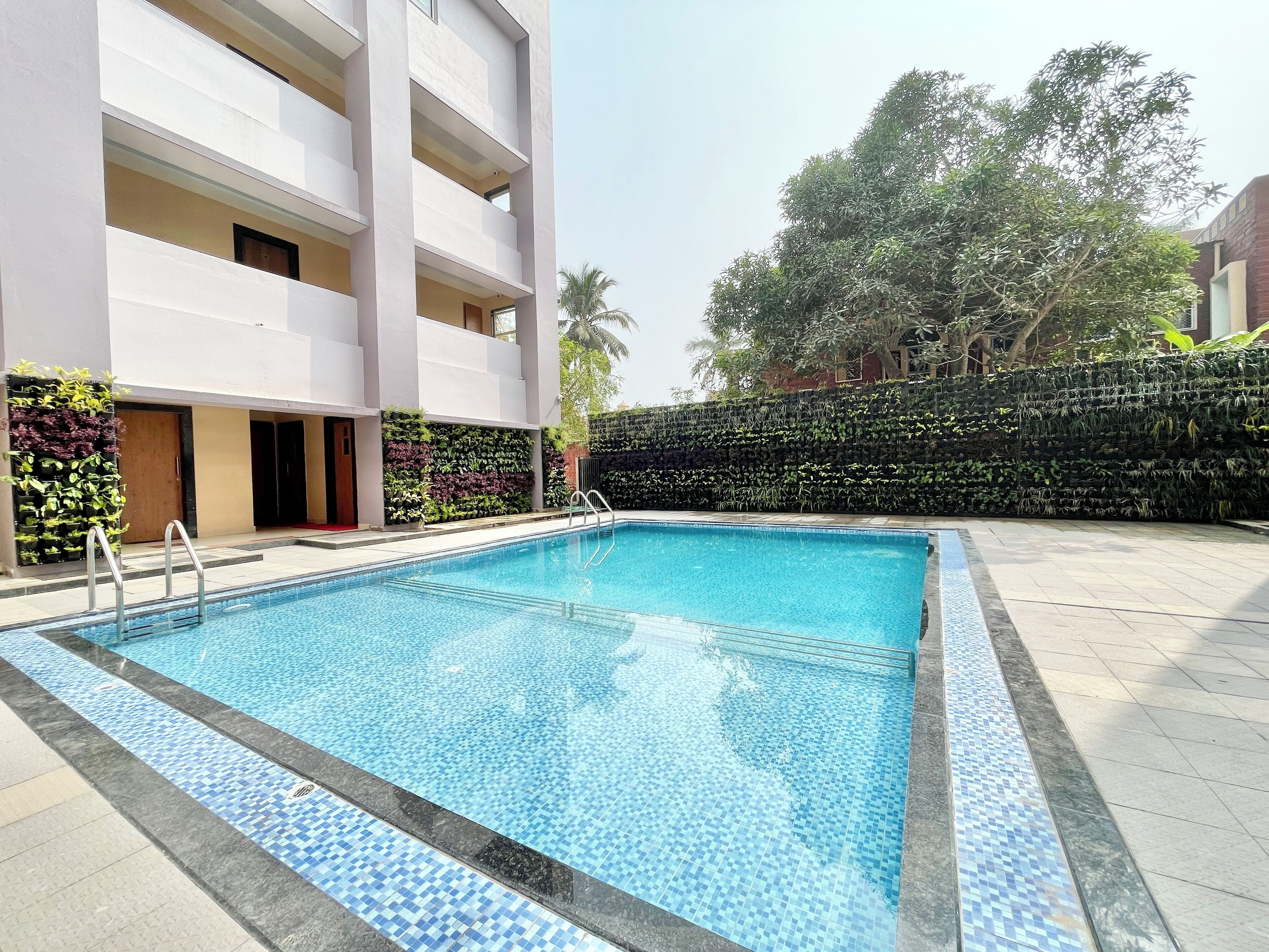 A swimming pool with a tiled edge, surrounded by a building with multiple balconies and a landscaped area with plants and a hedge.