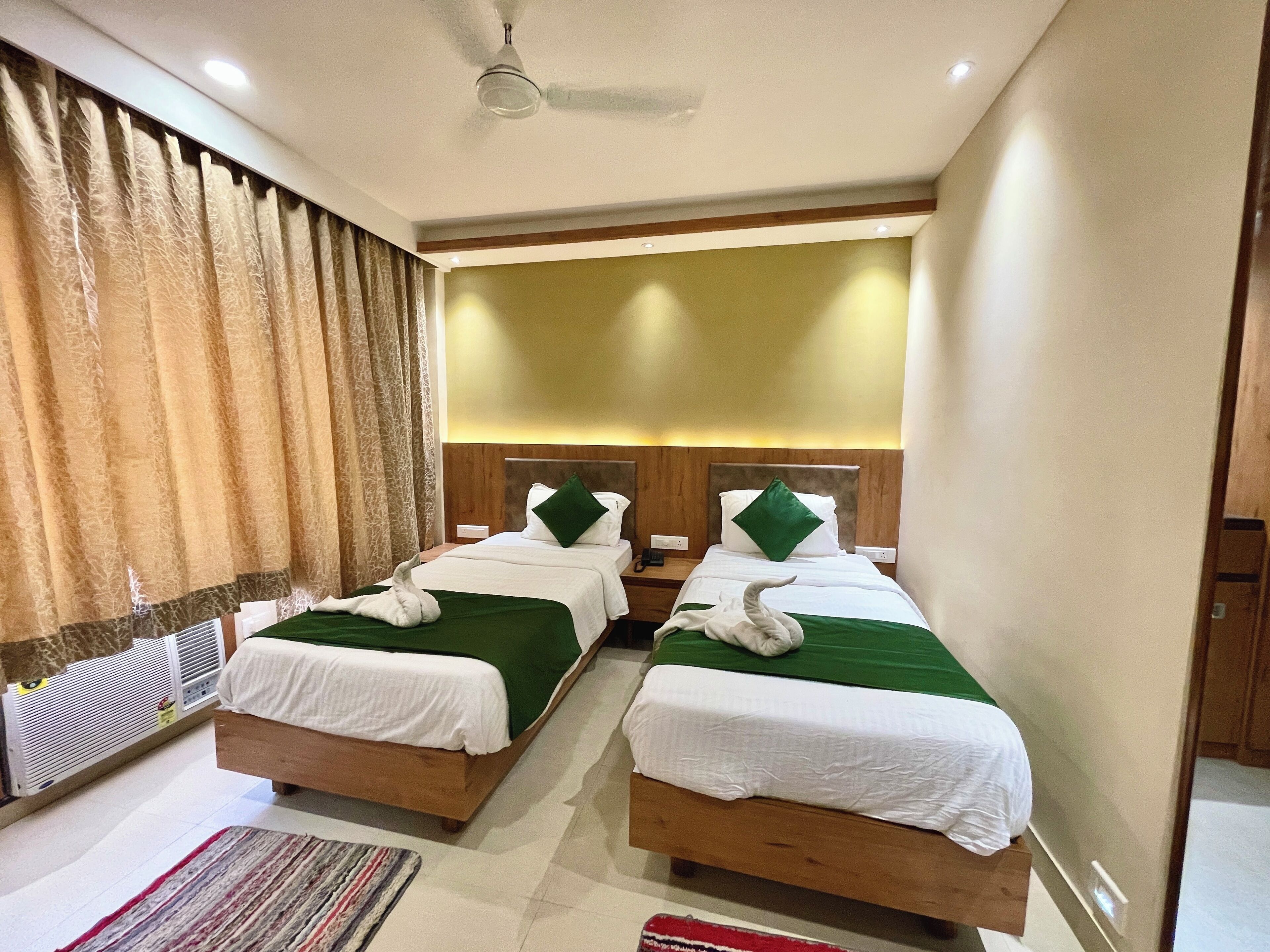 A hotel room with two beds, each with a green bedspread and white linens, a ceiling fan, and a large window with curtains.