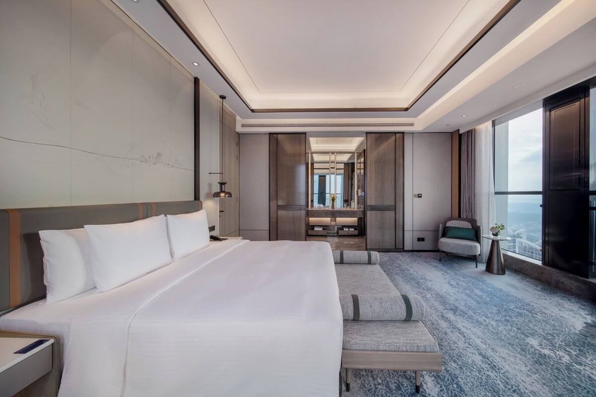A modern hotel room with a large bed, a seating area, a vanity with a mirror, and a view of the city.
