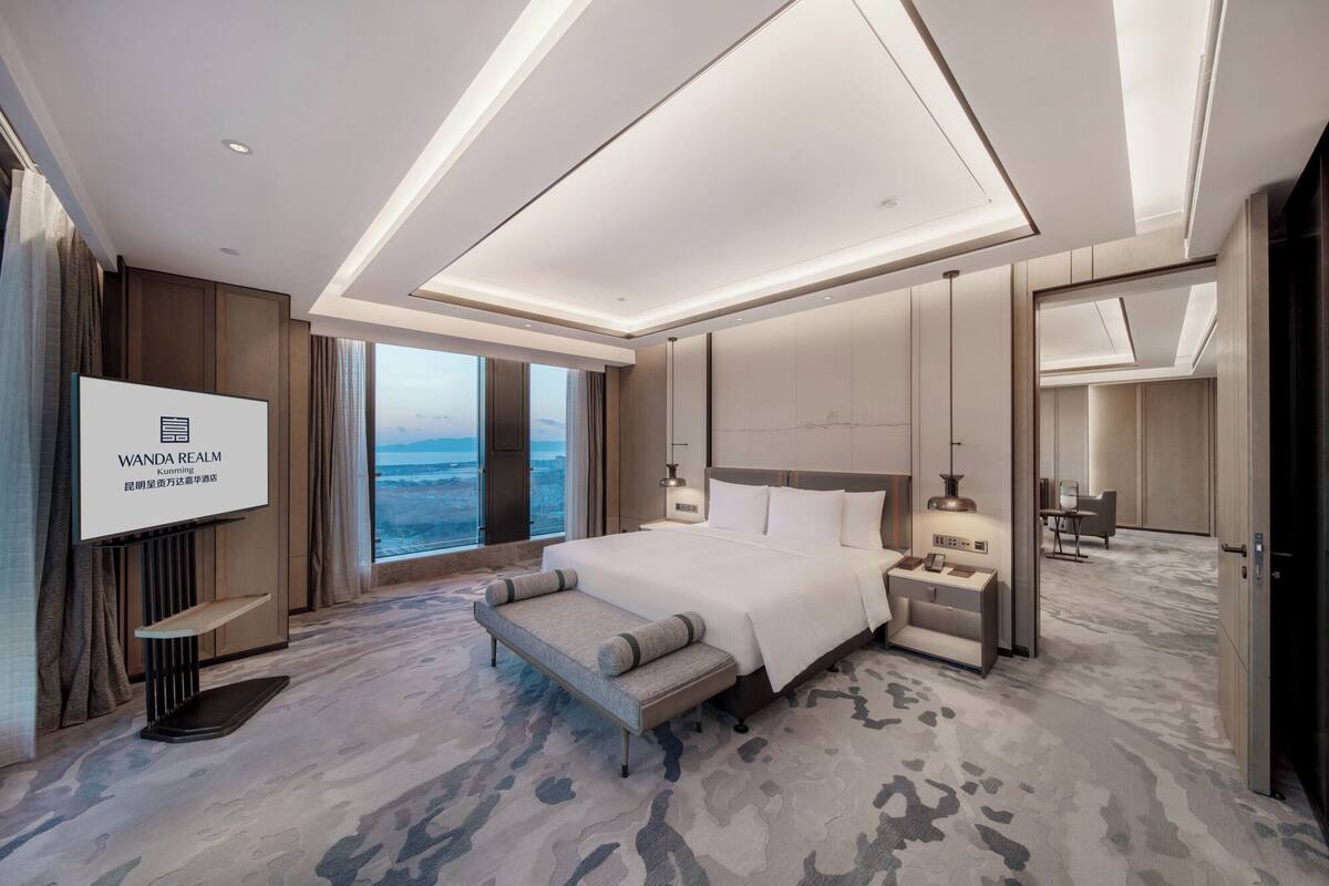 A modern hotel room with a large bed, bedside tables, a TV, and a view of the city.
