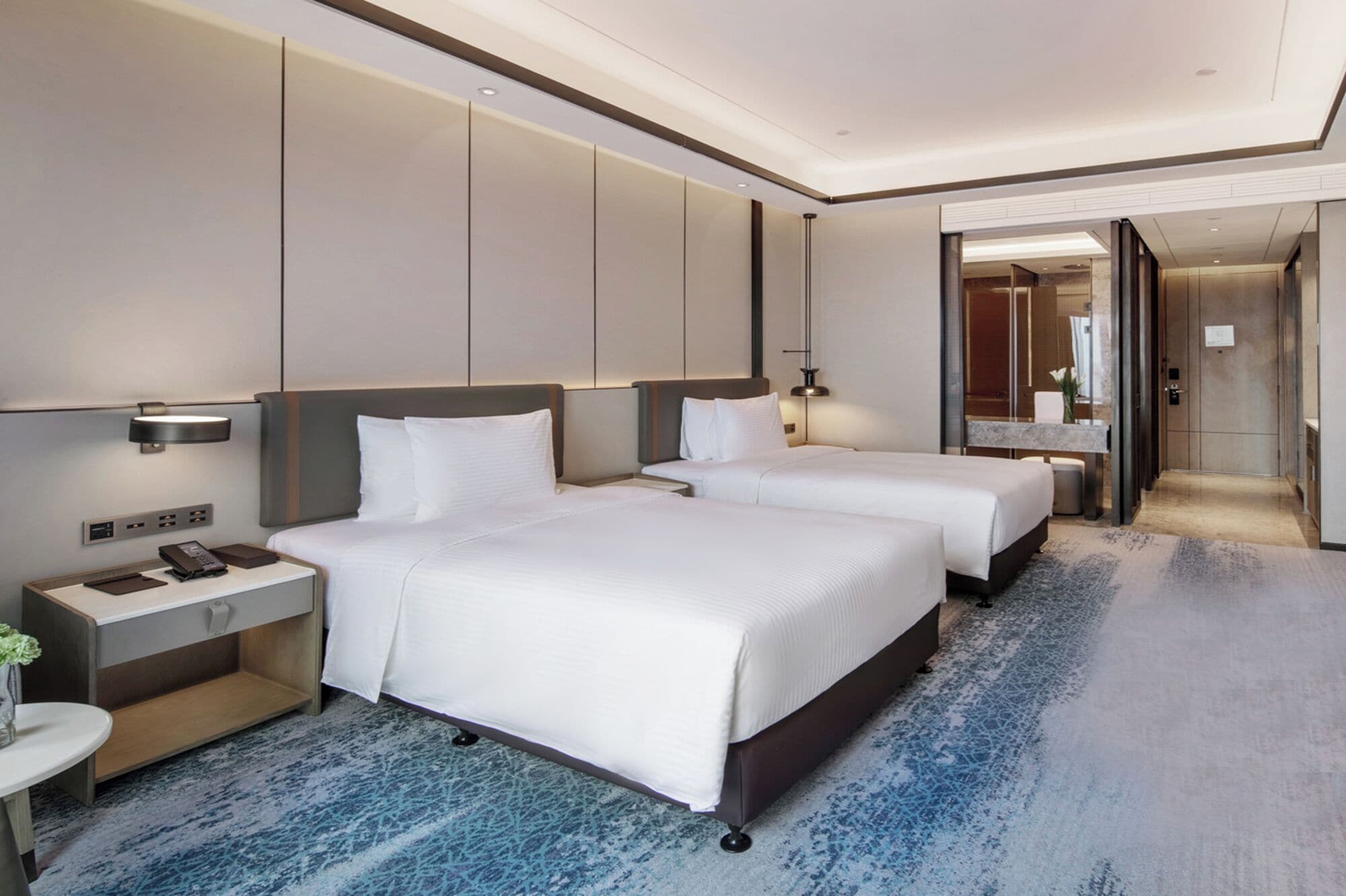 Habitación de hotel con dos camas, un escritorio con teléfono y lámpara, baño con espejo y lavamanos, y piso alfombrado.