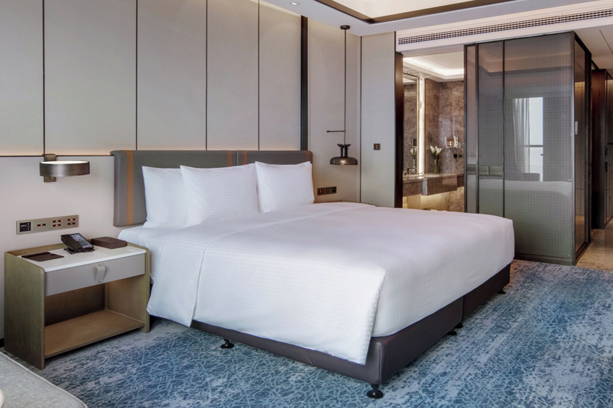 A modern hotel room with a large bed, a bedside table with a phone, a nightstand, a bathroom with a mirror, and a shower area.