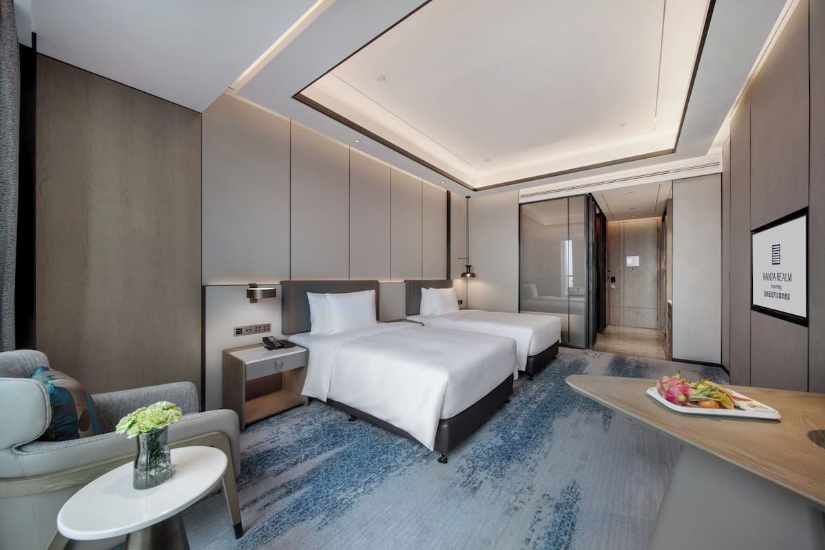 A modern hotel room with a large bed, two bedside tables, a seating area with a sofa, and a small table with a fruit plate.