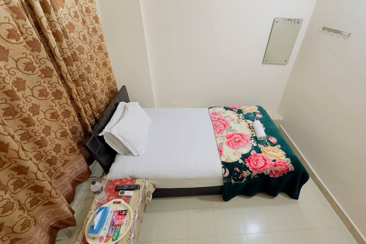 Una cama pequeña y bien hecha con una colcha estampada con flores, una mesita de noche con una botella y un control remoto, y una cortina con diseño de hojas.