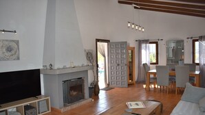Een woonkamer met een open haard, een televisie, een eethoek met stoelen en een keuken met een koelkast.