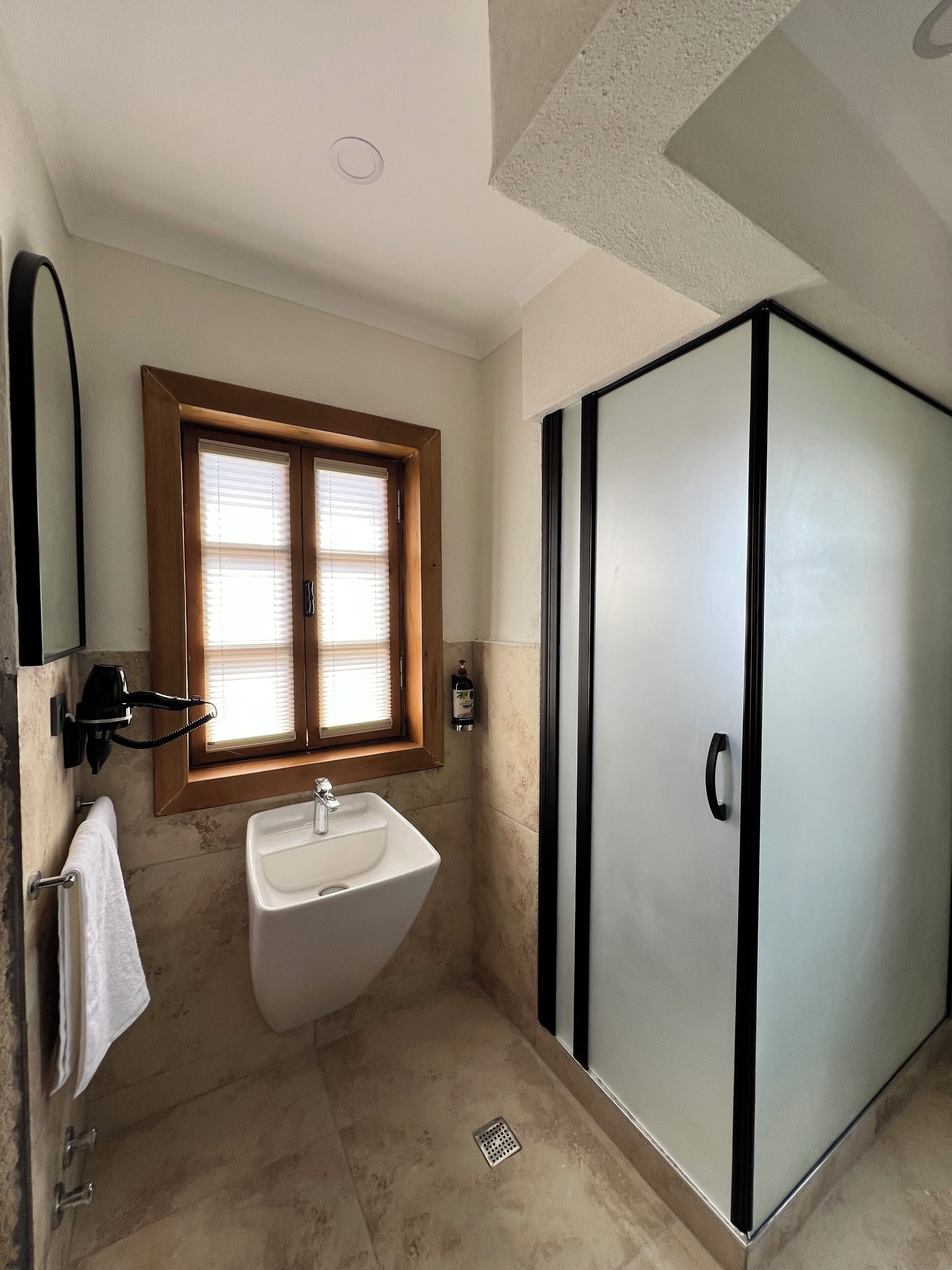 A bathroom with a sink, a window with blinds, a towel rack, and a shower area with a glass door.
