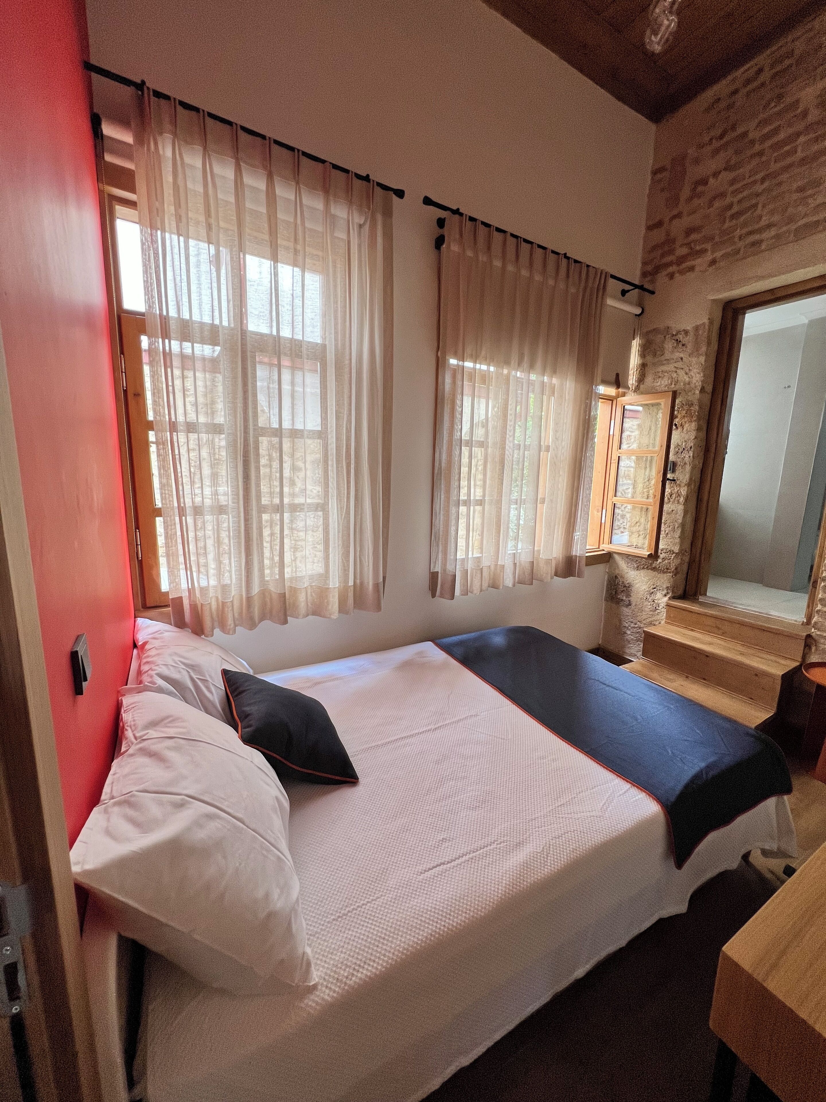 Ein Schlafzimmer mit einem Bett, einem Fenster mit Vorhängen, einer hölzernen Bank und einer Steinwand.