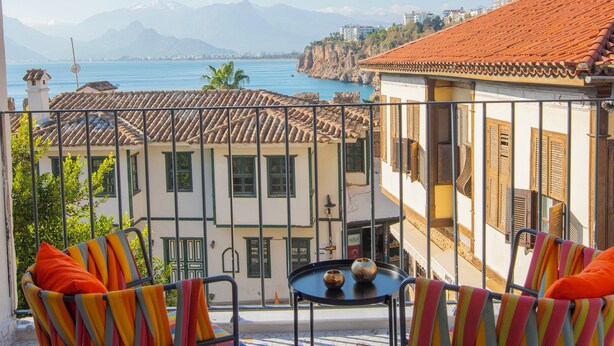 A balcony with two striped chairs and a small table, overlooking a coastal town with terracotta-roofed buildings and a clear blue sea. - Oldies Hotel Bistro Restaurant (Antalya)