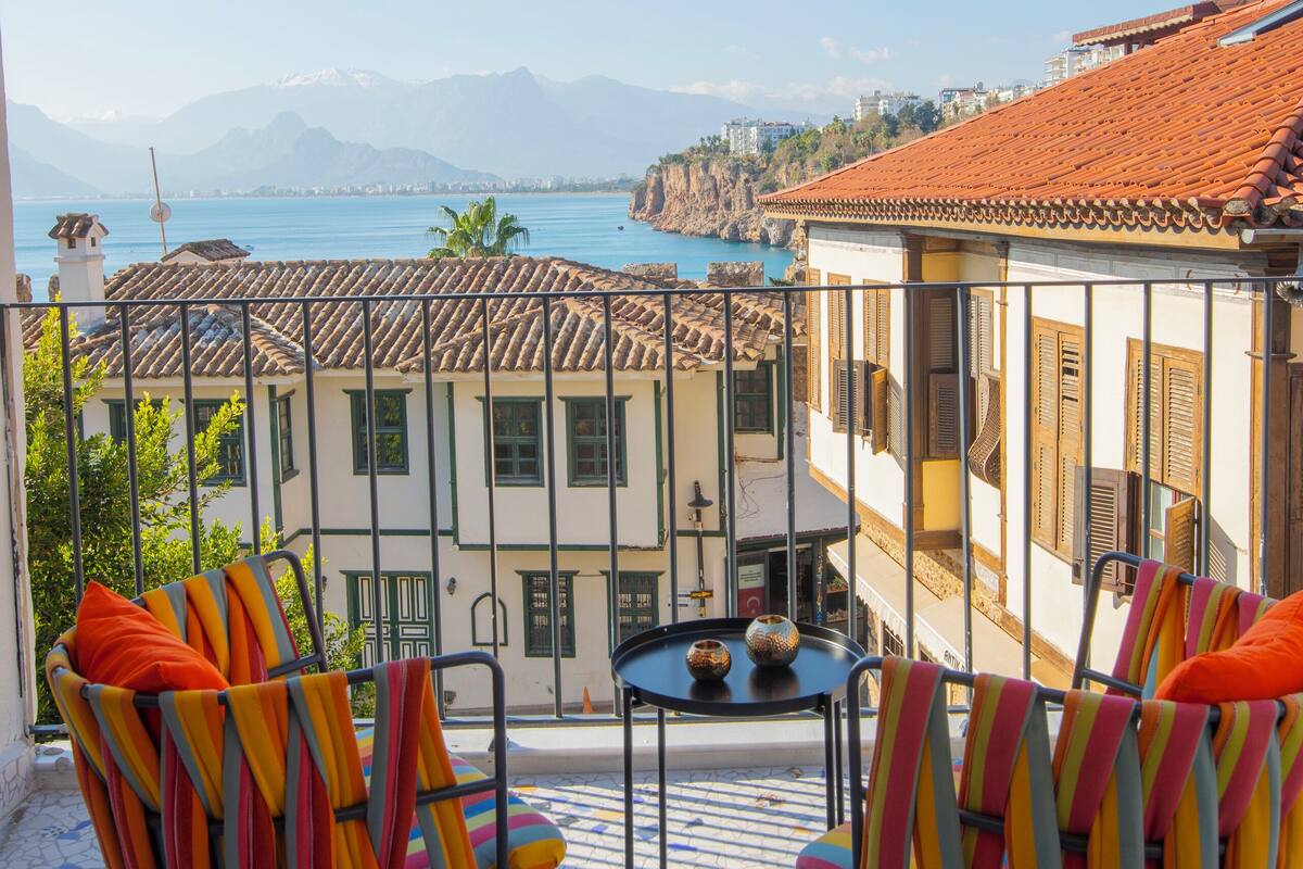 Un balcón con dos sillas a rayas y una mesita pequeña, con vistas a un pueblo costero de edificios con tejados de teja y un mar azul cristalino.