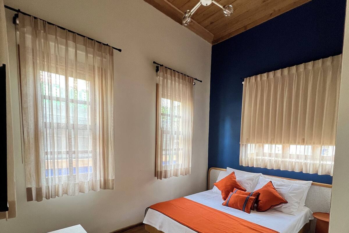 Un dormitorio con techo de madera, cama con ropa de cama naranja, dos ventanas con cortinas translúcidas y una mesa de madera.