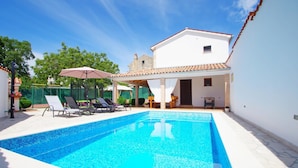 A rectangular swimming pool with clear blue water, surrounded by a white tiled deck. There are several lounge chairs and a covered seating area with a table and chairs. - Charming 3-bedroom house with private pool and beautiful garden (Šajini)