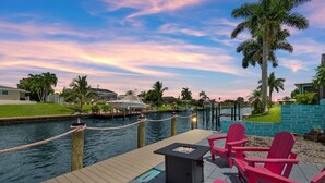 Eine Uferterrasse mit Feuerstelle und roten Adirondack-Stühlen, mit Blick auf einen Kanal, der von Palmen und Häusern im Hintergrund gesäumt ist.