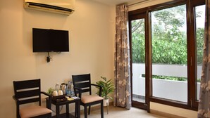 Una habitación con televisión de pantalla plana, dos sillones, una mesita con botellas y una planta, y un ventanal con cortinas.