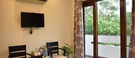 A room with a flat-screen TV, two chairs, a small table with bottles and a plant, and a large window with curtains.