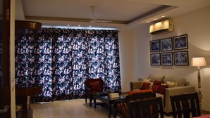 Sala de estar con sofá, sillones, una mesa de centro y televisor. Hay una cortina con estampado floral, un ventilador de techo y aire acondicionado instalado en la pared.
