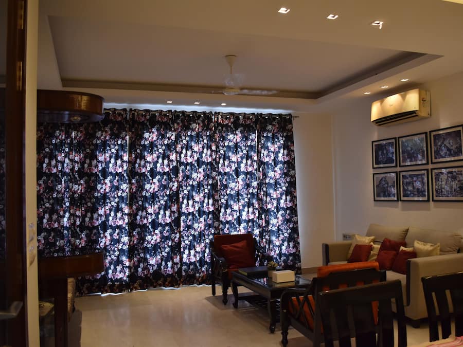Un salon comprenant un canapé, des fauteuils, une table basse et un téléviseur. On y trouve un rideau à motifs floraux, un ventilateur de plafond et un climatiseur fixé au mur.
