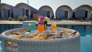 Desayuno junto a la piscina, con una cesta de mimbre que contiene diversos alimentos y bebidas, rodeado de edificios modernos con forma de cúpula y números.