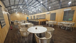 Un amplio comedor interior con mesas redondas y sillas de mimbre, un techo alto con vigas metálicas y paneles de madera en las paredes.