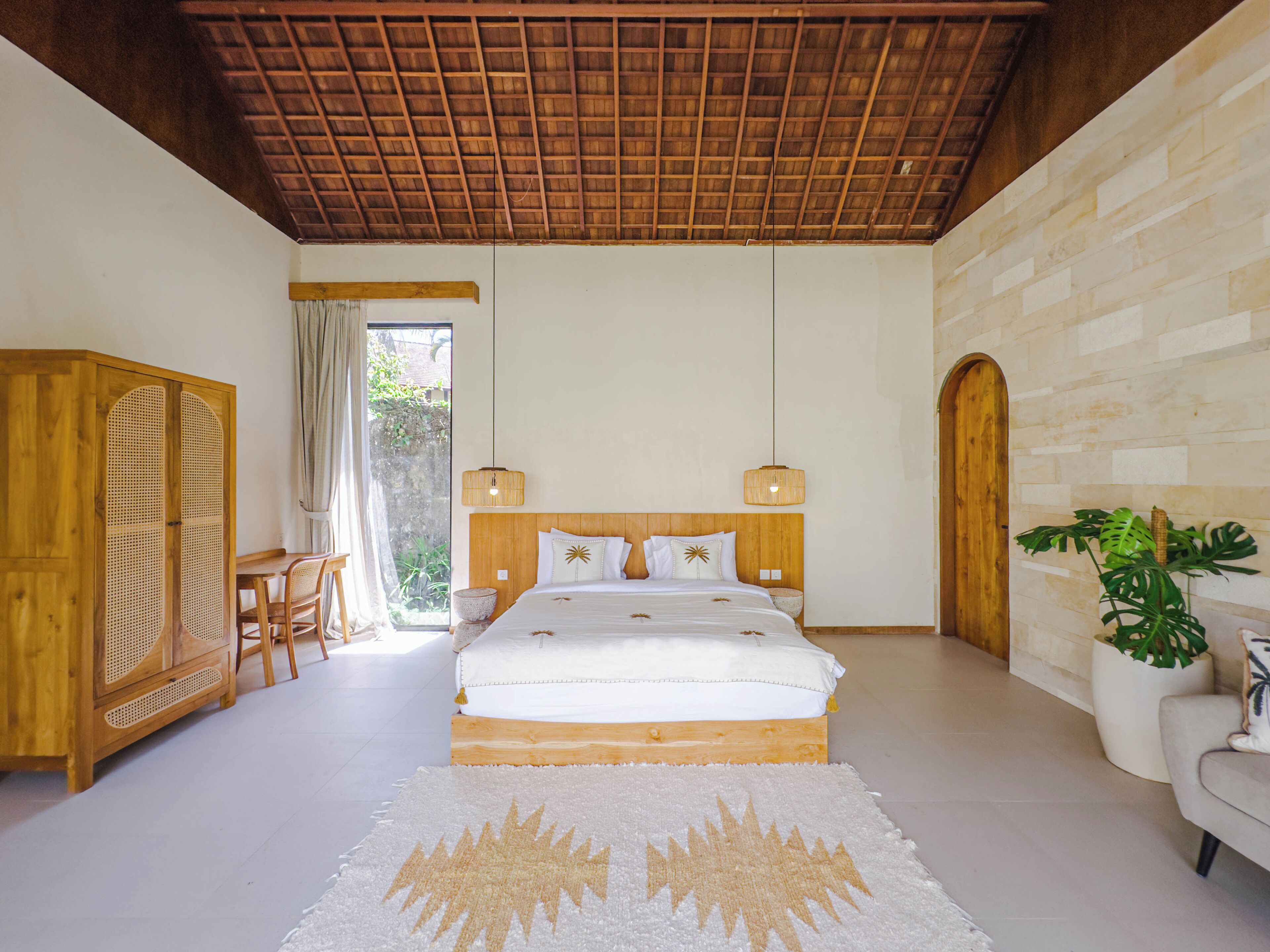 A bedroom with a wooden ceiling, a bed with white bedding, a wooden wardrobe, a dining table with chairs, and a potted plant.