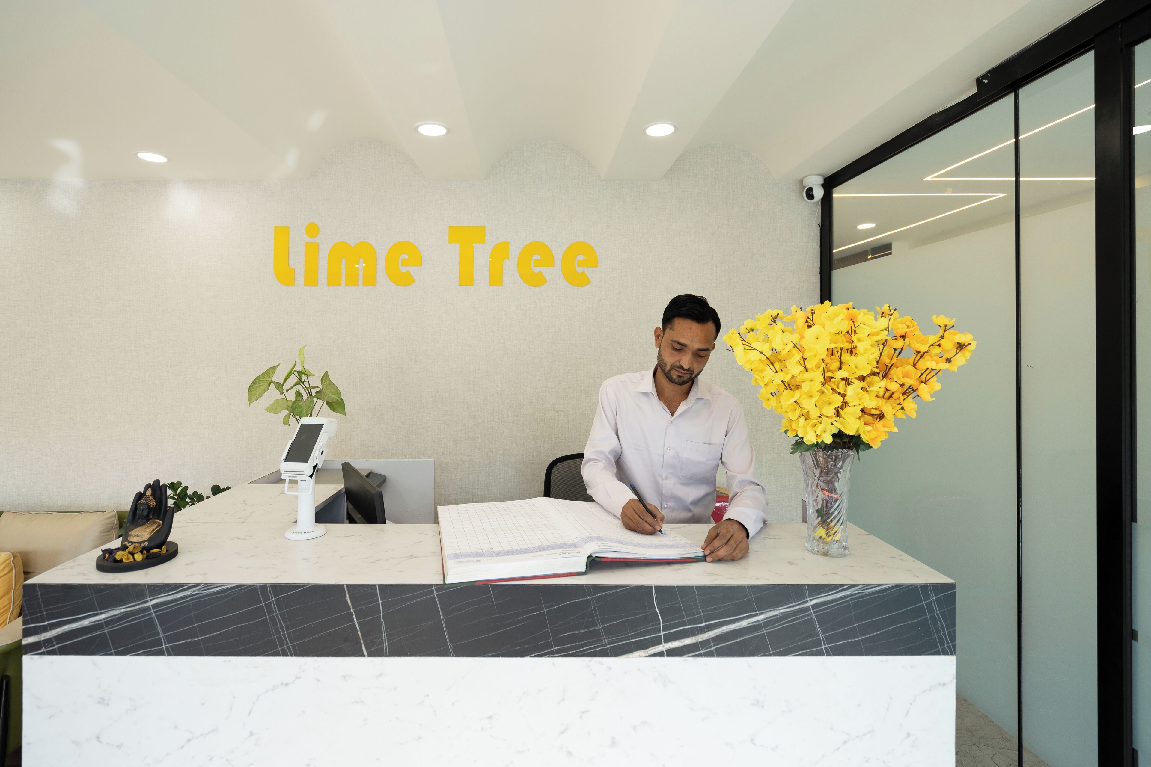Ein Empfangsbereich mit einem schreibenden Mann, einer Pflanze, einer Vase mit gelben Blumen und einem Schild mit der Aufschrift 'lime Tree'.